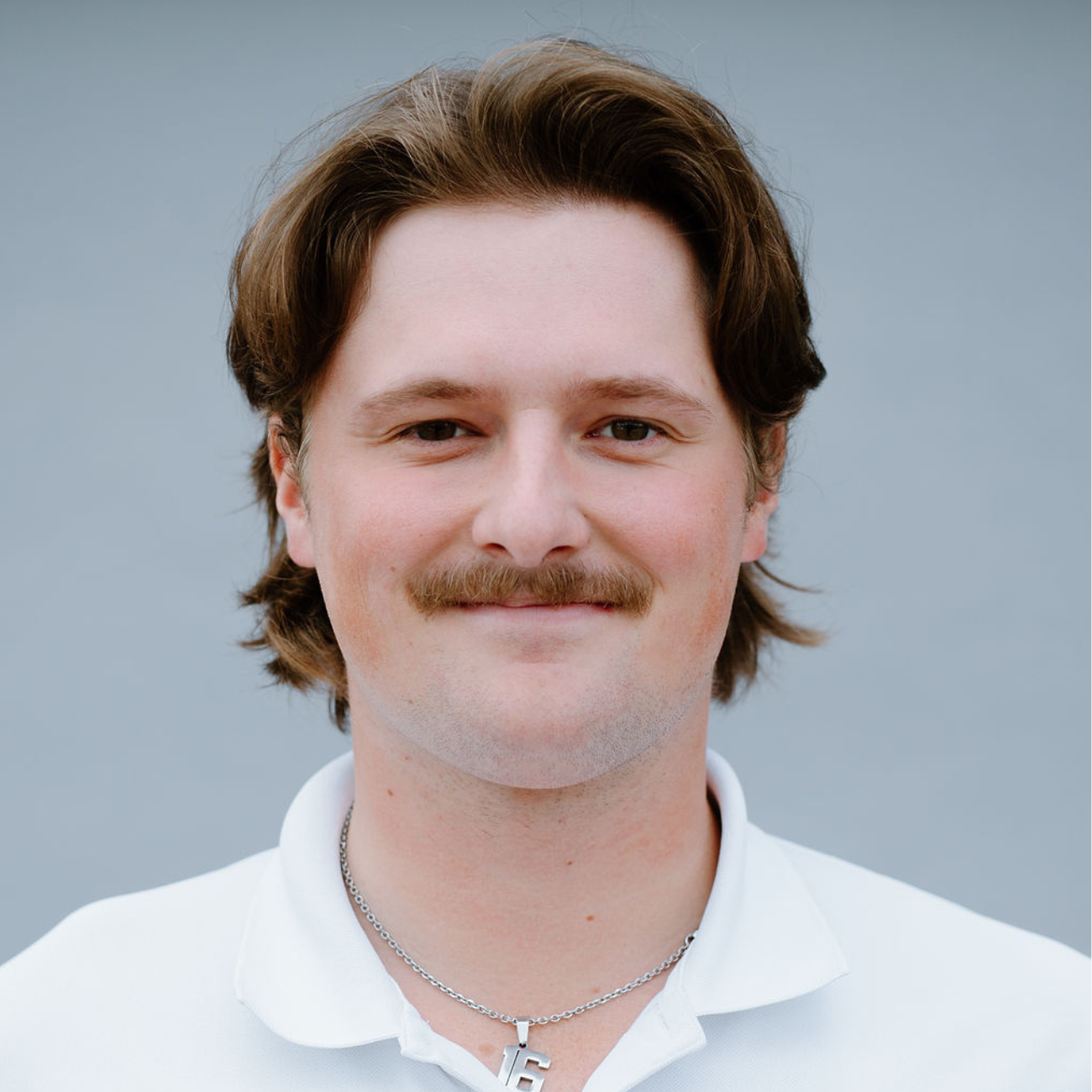 Man with medium-length brown hair and a mustache, wearing a white collared shirt and a silver necklace, smiling at the camera against a plain gray background.