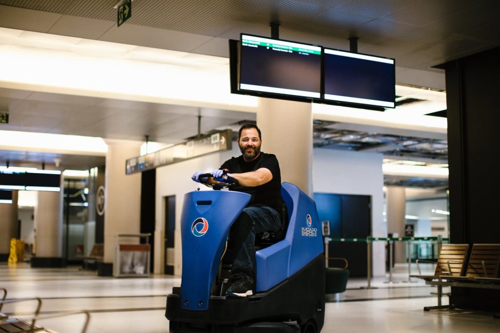 A man rides a blue floor cleaning machine inside a spacious, modern building with empty benches and digital screens overhead.