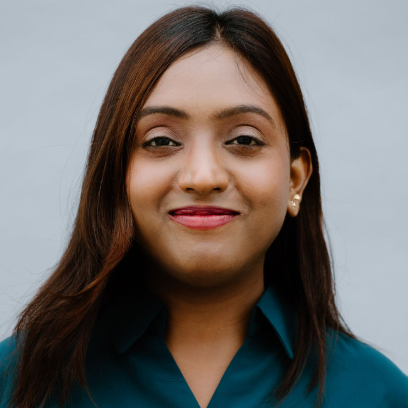A woman with medium brown hair wearing a dark teal collared shirt smiles softly at the camera against a plain light gray background.