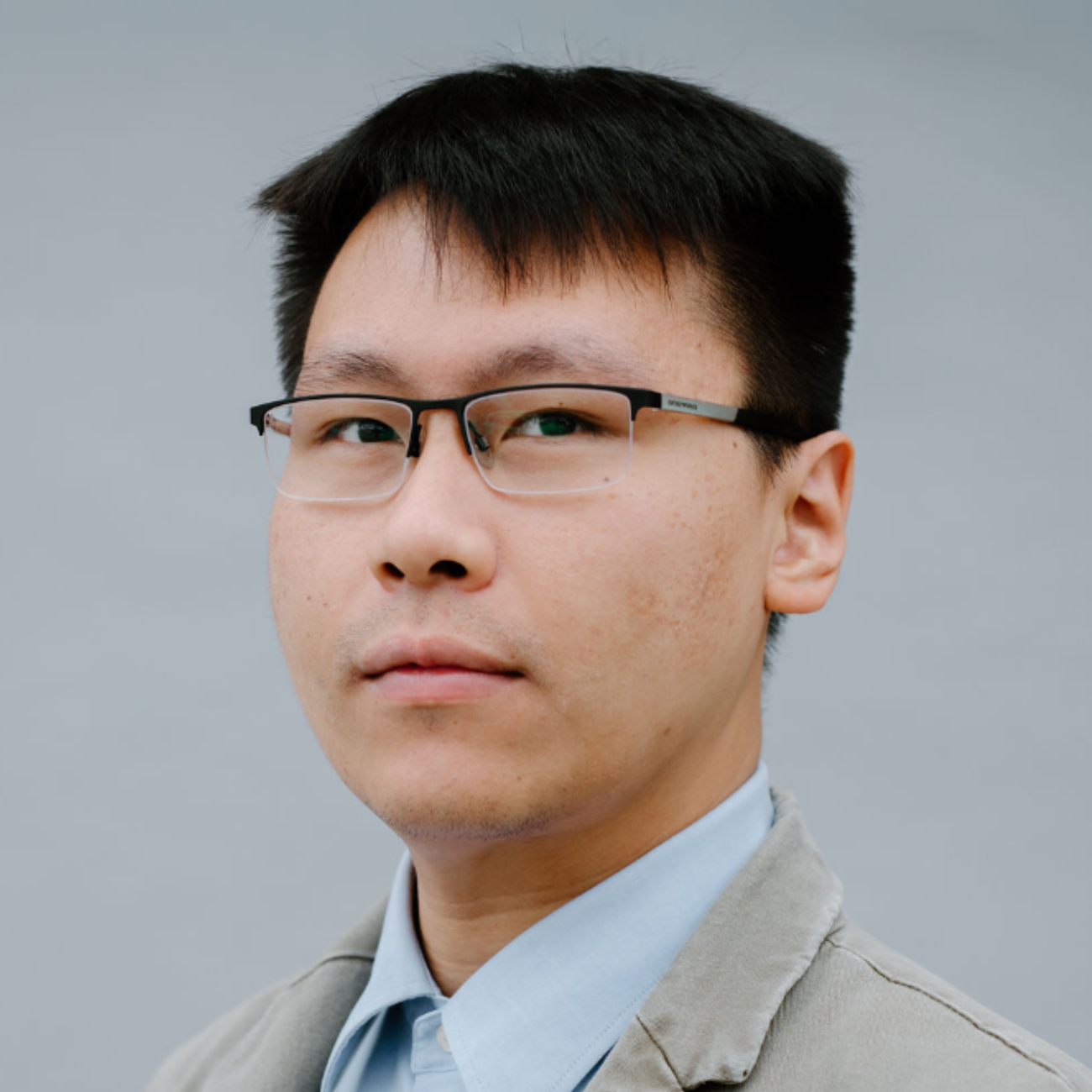 A man with short black hair and glasses wears a light blue collared shirt and a gray blazer, posing in front of a plain light gray background.