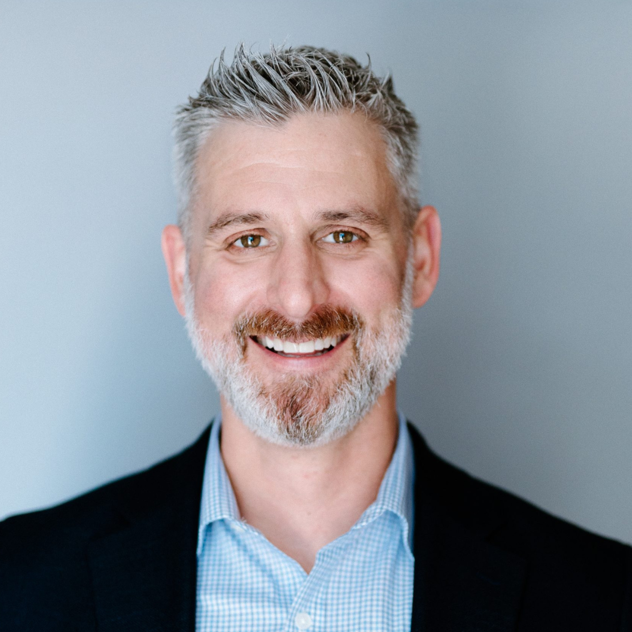 Smiling man with short gray hair and a beard, wearing a blue checkered shirt and dark blazer, standing against a light gray background.